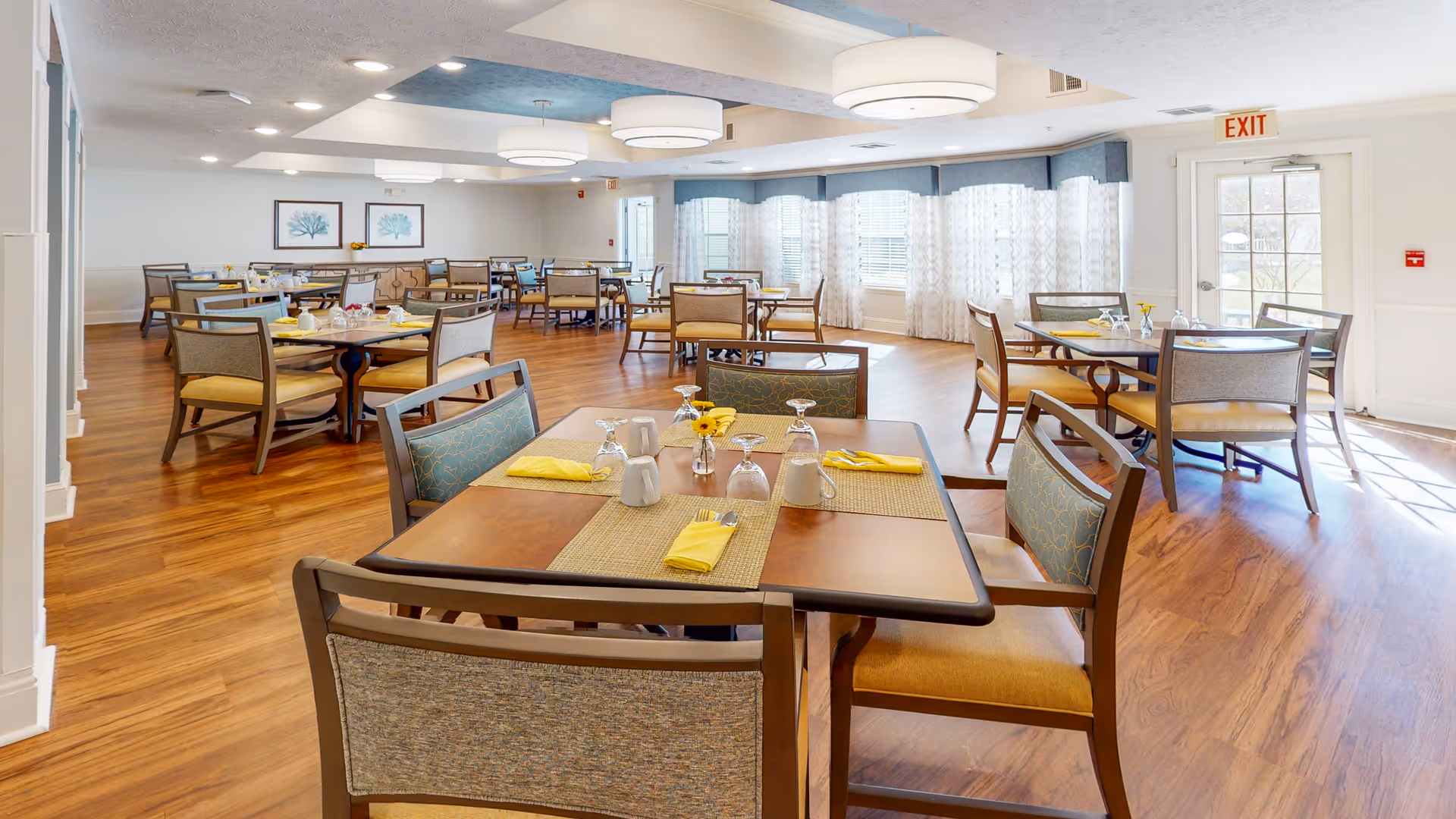 A bright and spacious dining room with multiple wooden tables and chairs arranged neatly. Each table is set with placemats, yellow napkins, glassware, and small flower vases. Large windows with sheer white curtains allow natural light to fill the room, and modern ceiling lights hang overhead. The floor is a warm wood tone, and framed artwork is visible on the far wall.