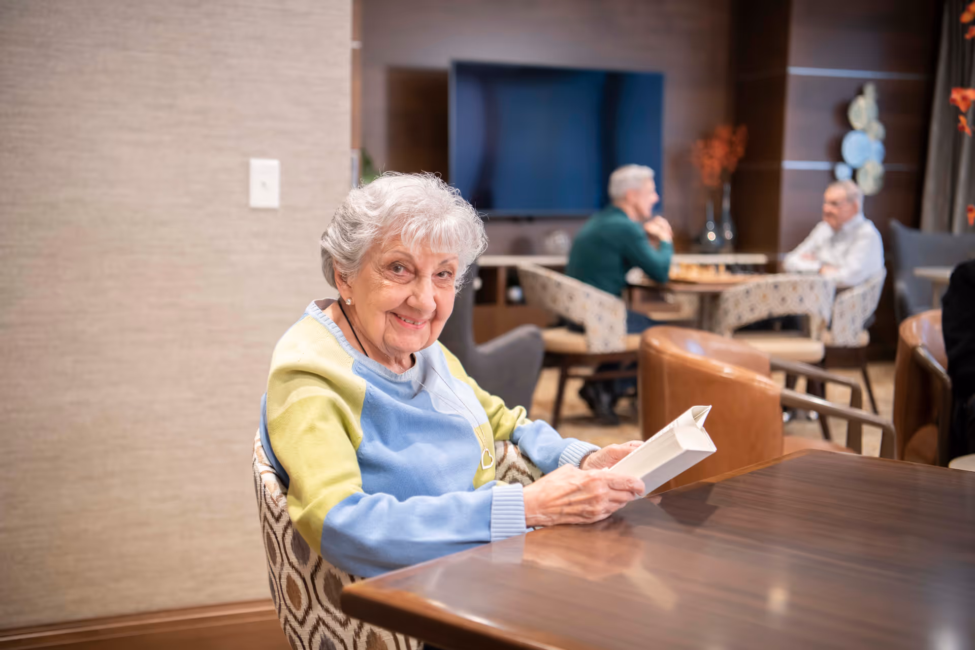 An elderly woman with gray hair wearing a blue and yellow sweater is sitting at a wooden table reading a book in a cozy common area. In the background, two elderly men are sitting at another table playing a board game, with a large flat-screen TV mounted on the wall behind them.