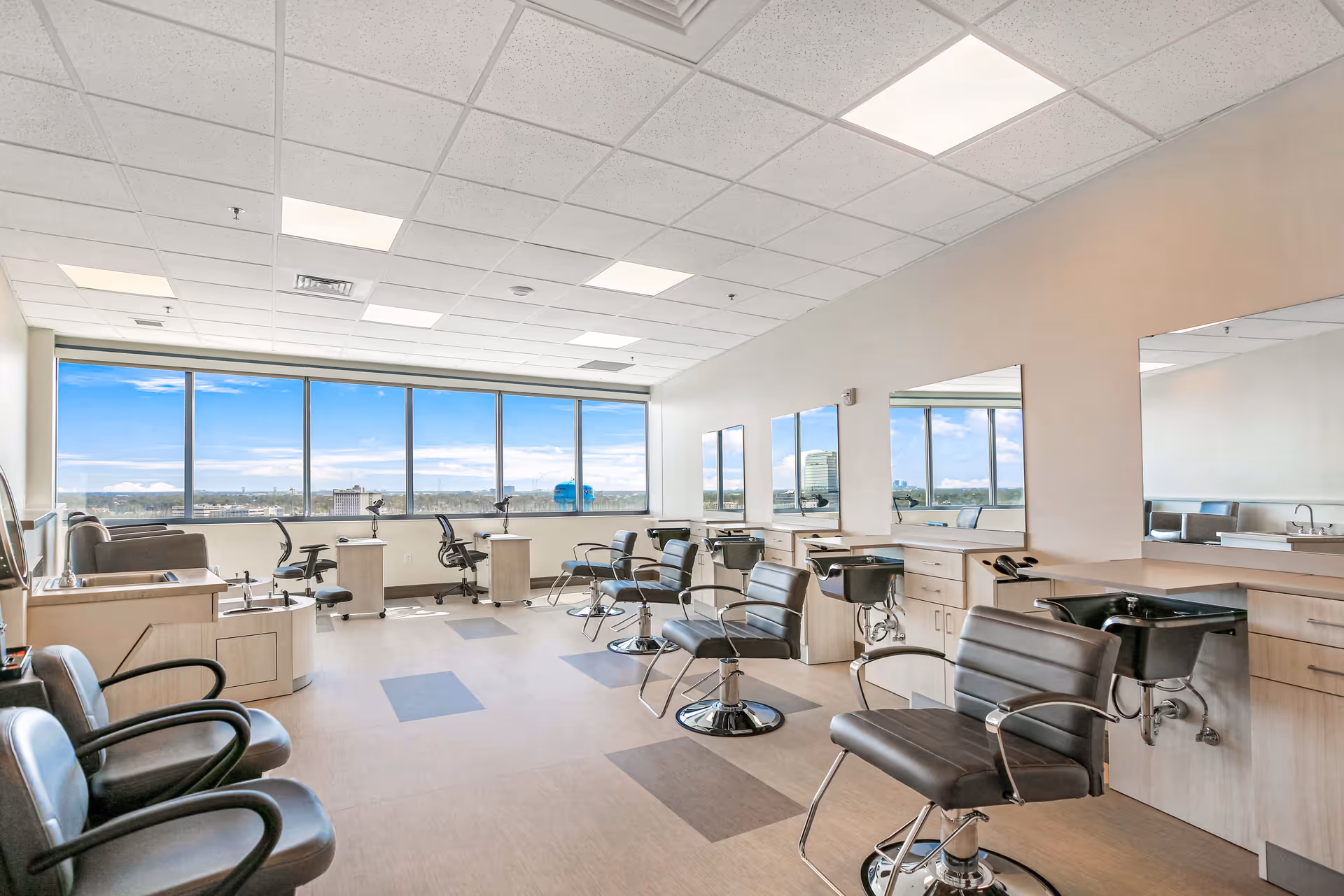 A bright and spacious salon area with multiple black salon chairs facing large mirrors mounted on the wall. The room has large windows offering a panoramic view of the city skyline under a blue sky with some clouds. The floor has a light-colored pattern with darker square accents, and the ceiling is fitted with recessed lighting panels.