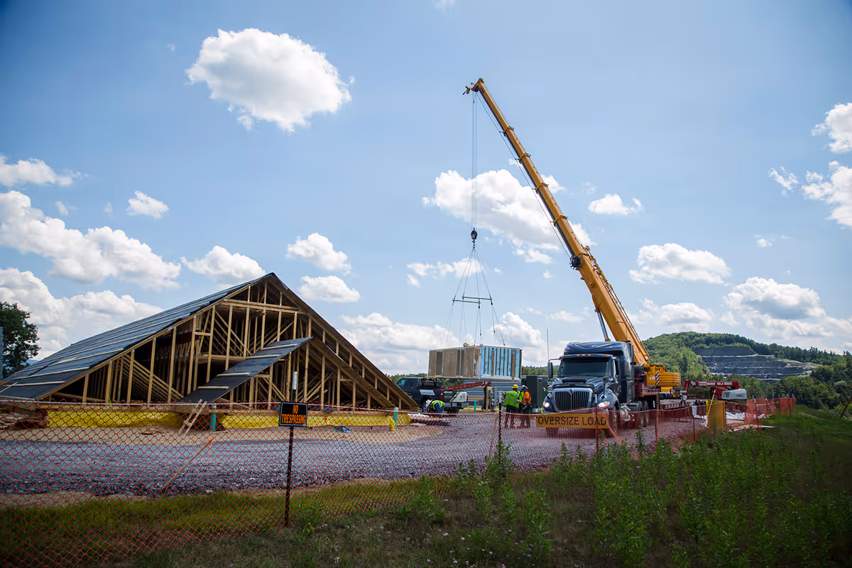 Construction site with a large crane lifting a prefabricated panel beside a wooden framed structure behind orange safety fencing under a blue sky.