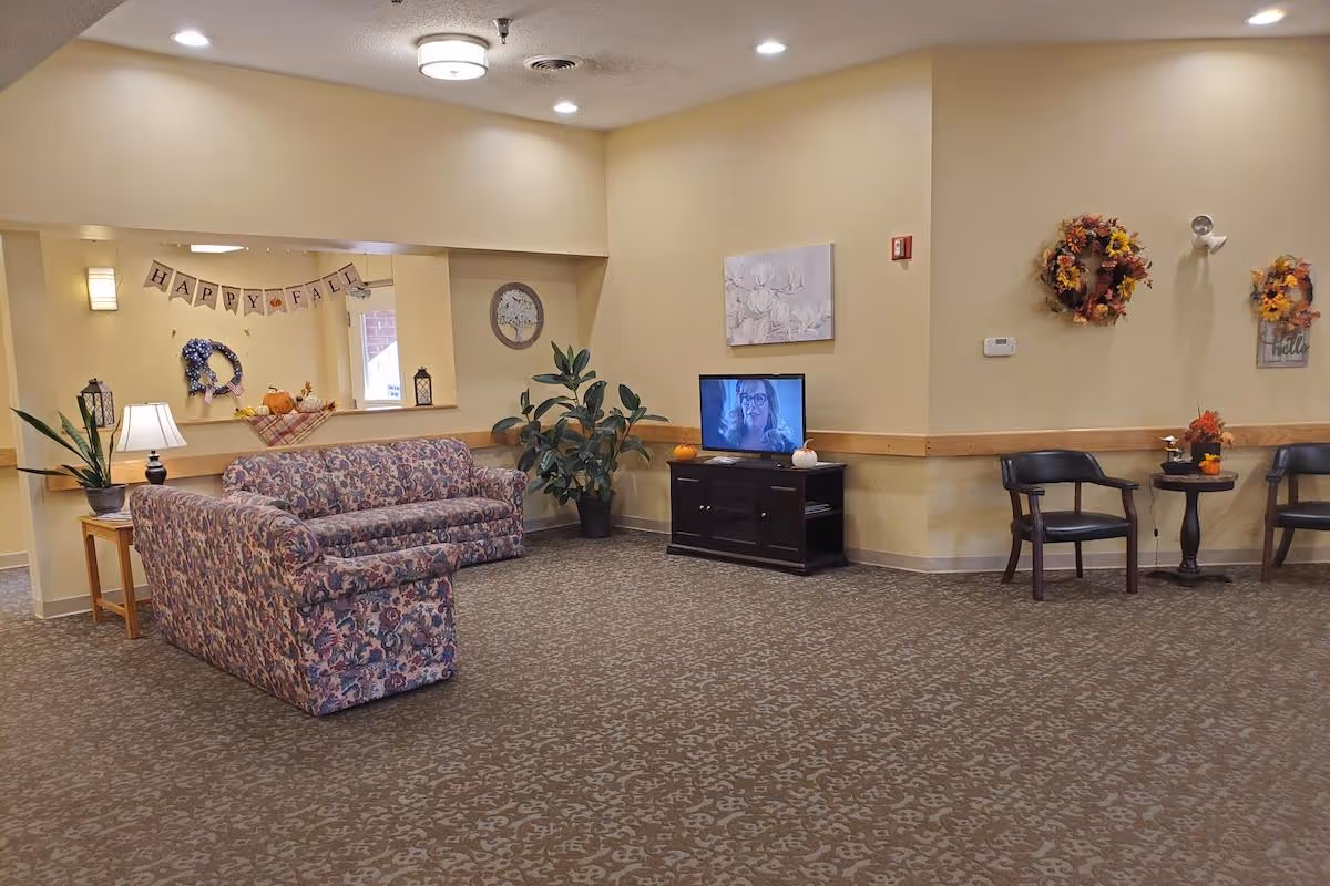 A cozy living room area in a senior living facility decorated for fall with a floral patterned sofa, a TV on a wooden stand, two black chairs with a small round table between them, fall wreaths on the wall, and a 'Happy Fall' banner reflected in a mirror.
