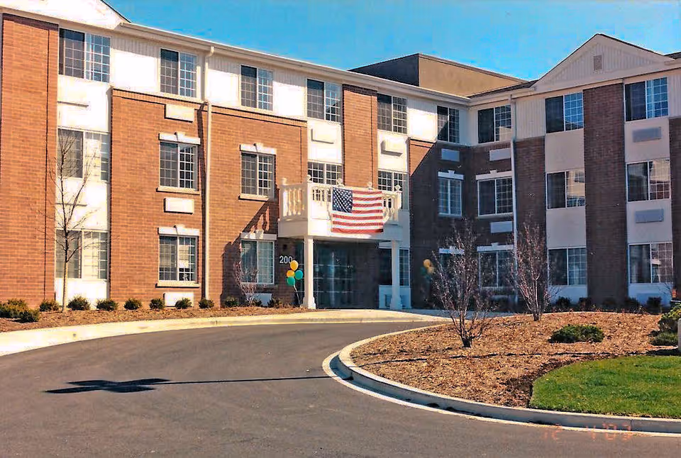 Exterior view of a three-story senior living apartment building with brick and white paneling. There is a curved driveway leading to the entrance, which has an American flag hanging above it and some balloons near the door. The landscaping includes small bushes and trees with mulch beds.