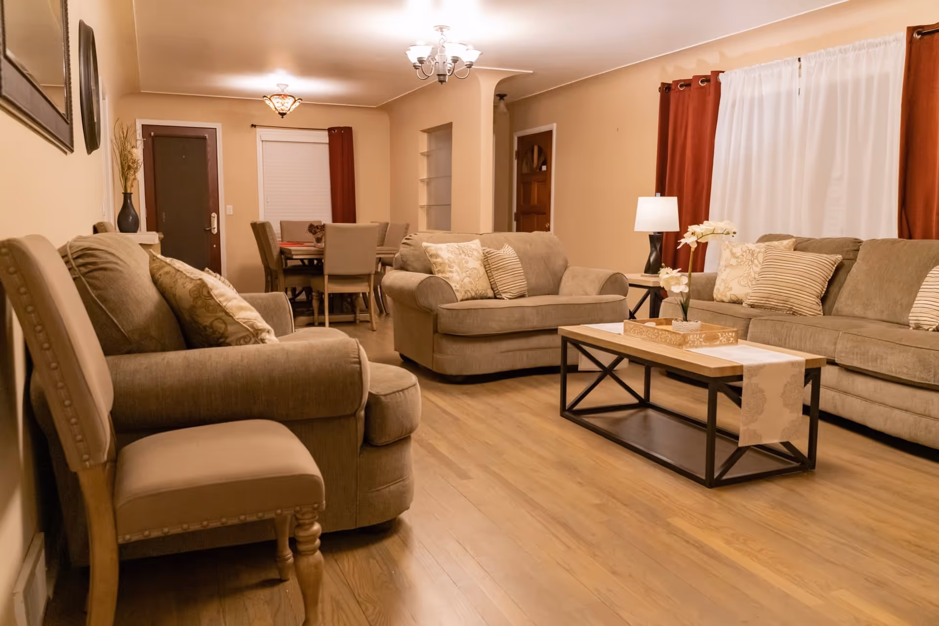 A cozy living room with beige walls and wooden flooring, featuring a beige sofa set with patterned cushions, a wooden coffee table with a decorative runner and flowers, a side table with a lamp, and a dining area with a table and chairs in the background. The room has red curtains and white blinds on the windows.