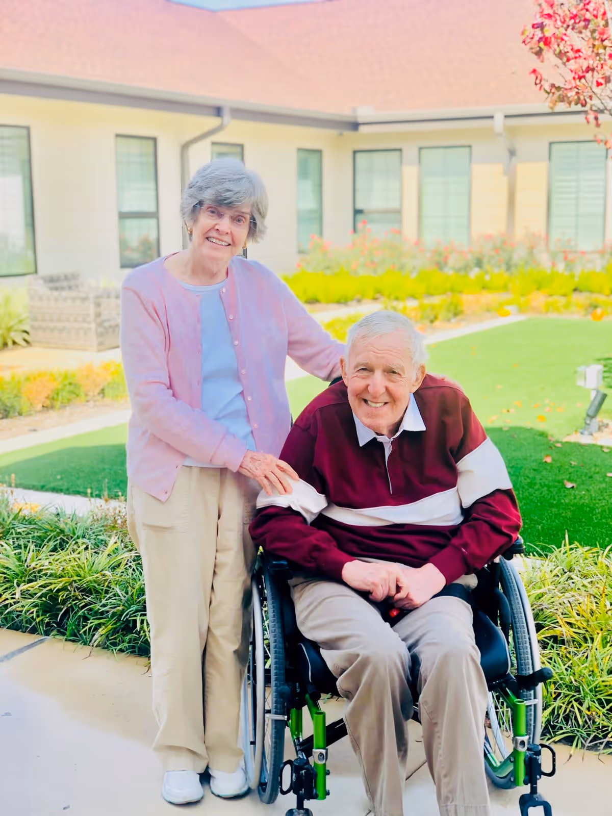 An elderly woman standing next to an elderly man seated in a wheelchair outside in a garden area of a senior living facility. The woman is smiling and has her hand on the man's shoulder. The man is also smiling. Behind them is a building with multiple windows and a well-maintained garden with green grass and flowering plants.