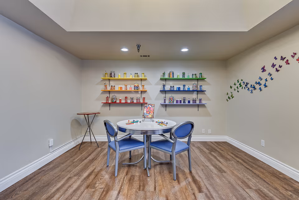 A small activity room with a round table and two blue chairs. On the table are colorful game pieces and a board. The back wall has shelves with jars filled with colorful items arranged by color, and a wall decoration of butterflies in a rainbow pattern on the right side.