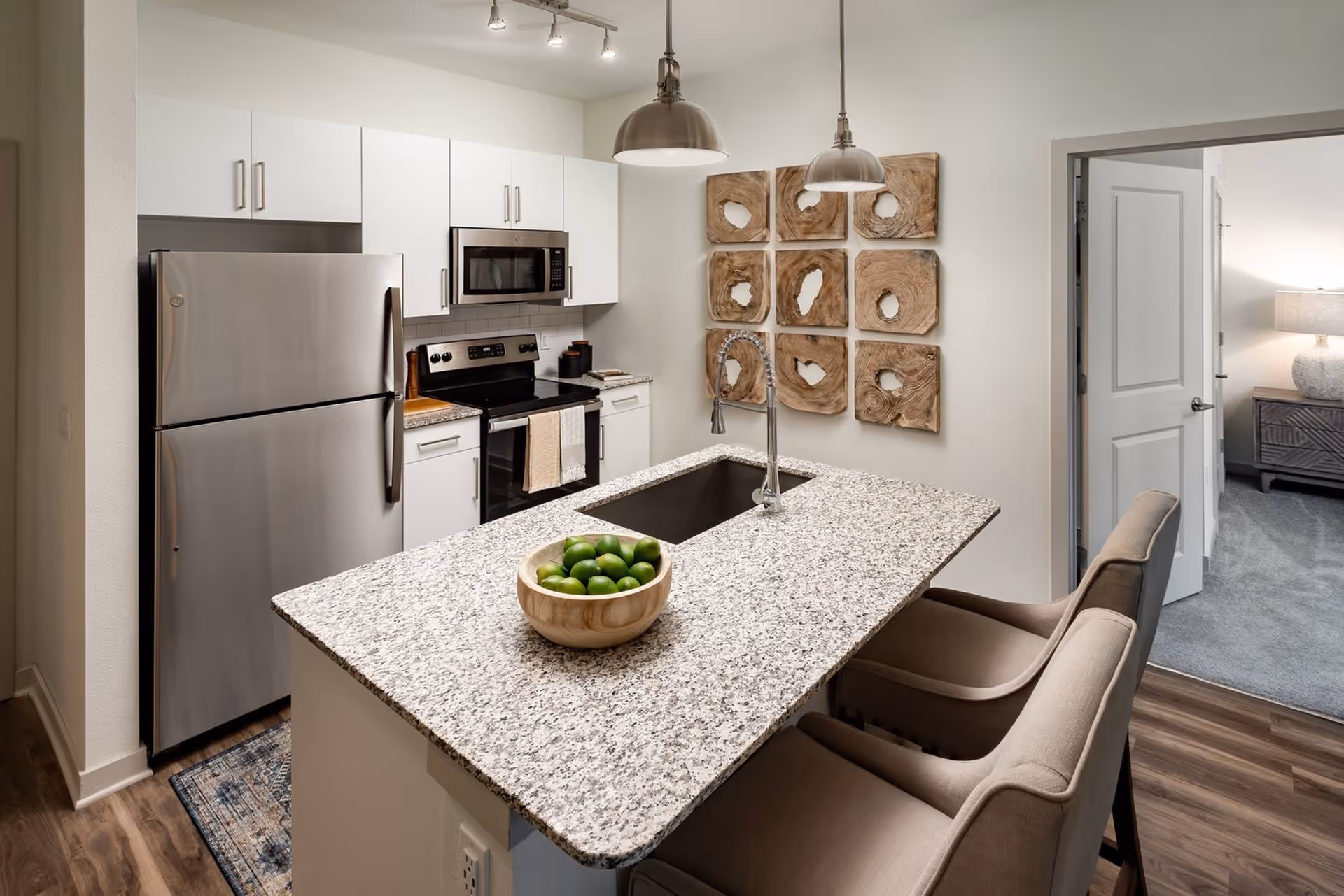 Modern open kitchen with a granite island and sink, stainless steel refrigerator and stove, two bar stools, decorative wall art, and a doorway leading to a bedroom.
