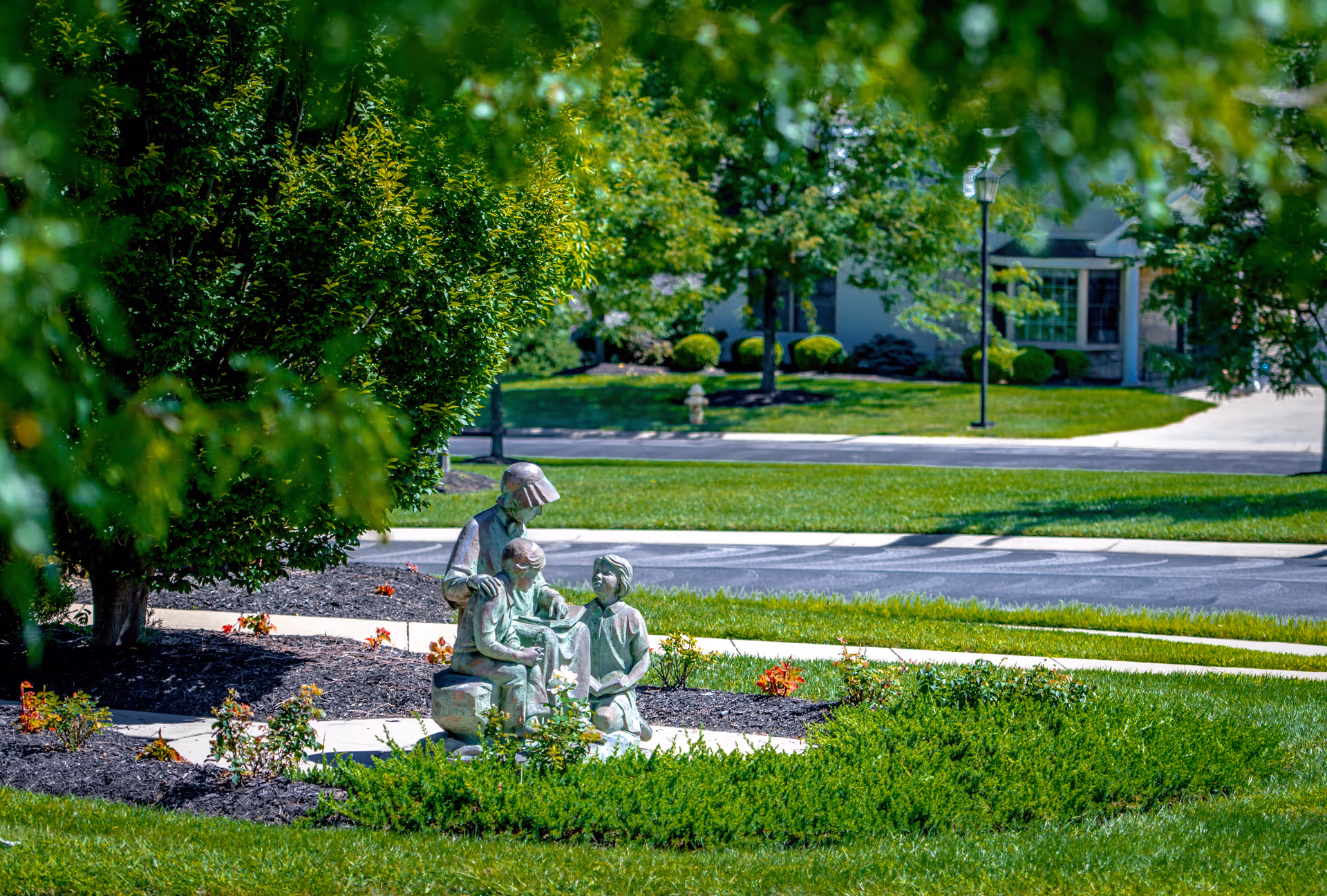 A garden area with a statue of three children sitting and interacting, surrounded by green bushes, trees, and a well-maintained lawn with a paved walkway and a building in the background.