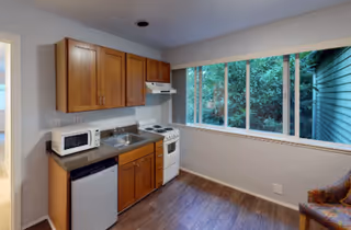 Small kitchenette with wooden cabinets, sink, microwave, mini-fridge, stove, and a large window overlooking greenery.
