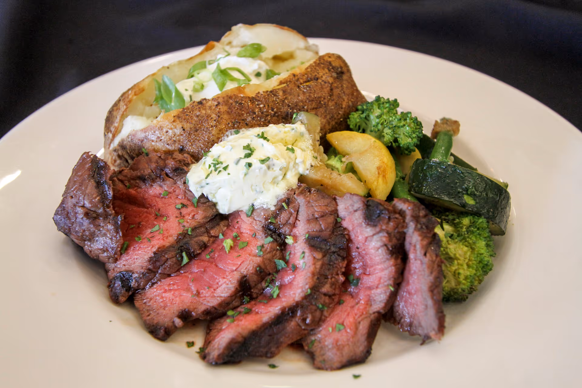 A plate of food featuring sliced medium-rare steak garnished with herbs, a baked potato topped with sour cream and green onions, and a side of steamed mixed vegetables including broccoli, zucchini, and yellow squash.