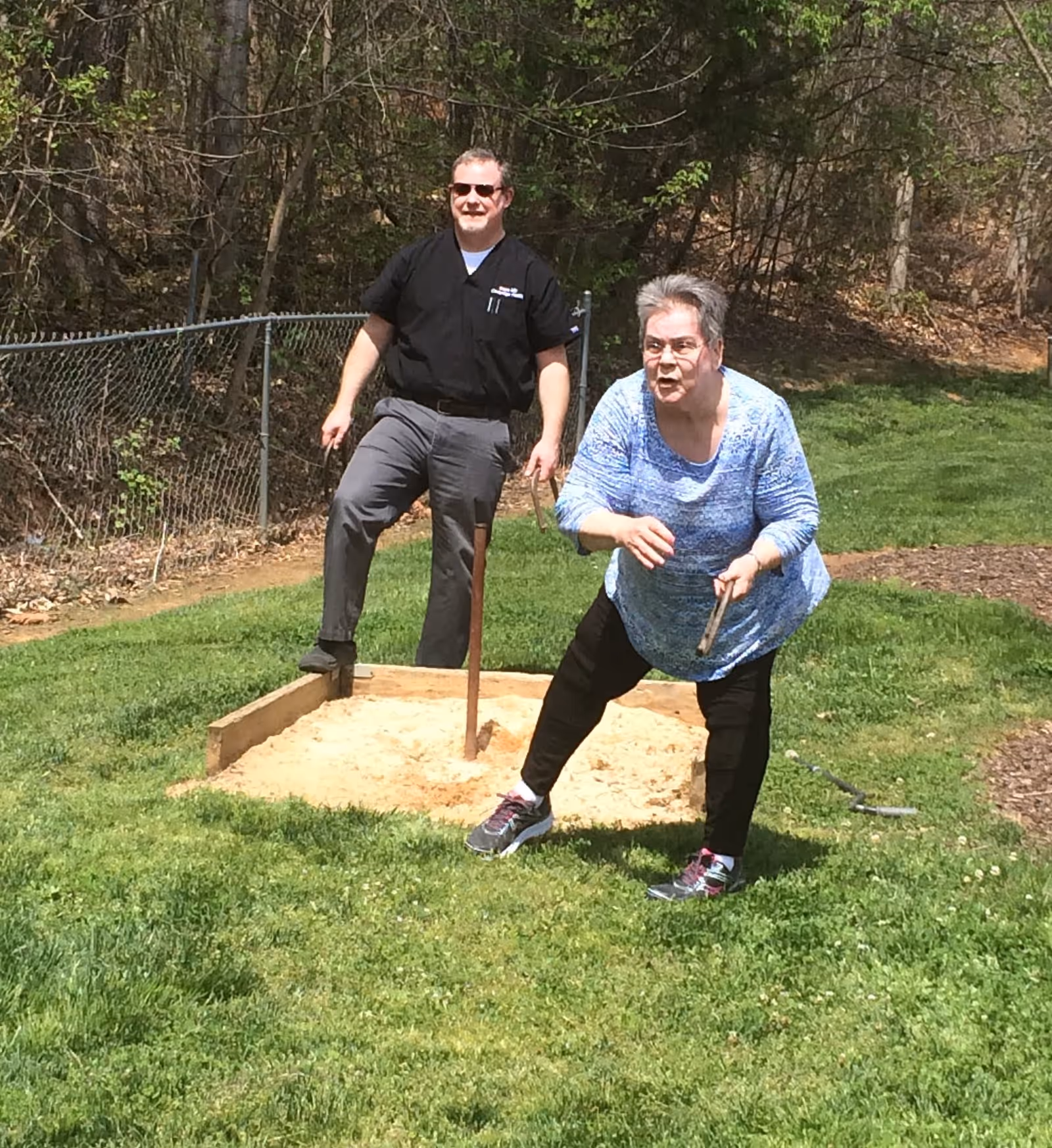 An elderly woman and a man are playing horseshoes outdoors on a grassy area with trees and a chain-link fence in the background. The woman is in the process of throwing a horseshoe while the man watches and smiles.