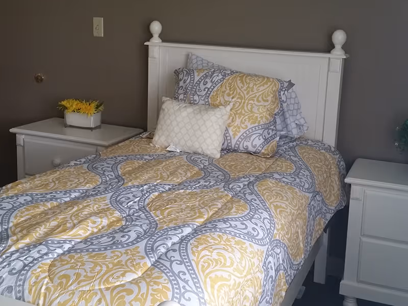 A neatly made single bed with a white wooden headboard and a patterned yellow, gray, and white comforter. There are two pillows, one with a matching pattern and one smaller white pillow. On each side of the bed are white nightstands, one with a small rectangular vase holding yellow flowers. The walls are painted gray.