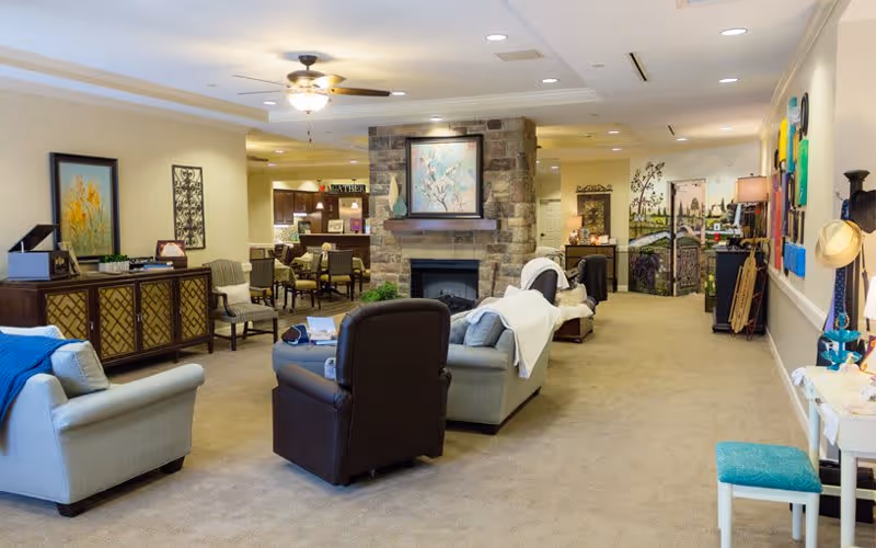 A spacious and well-lit living room area in a senior living facility featuring comfortable armchairs and sofas arranged around a stone fireplace with a painting above it. The room has beige walls and carpet, decorative artwork on the walls, and a sideboard with plants and decor. In the background, there is a dining area with tables and chairs, and a hallway leading to other rooms.