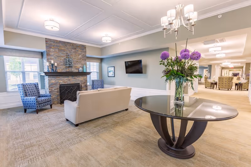 A bright and spacious living room area in a senior living facility featuring a beige sofa, two patterned blue armchairs, a stone fireplace with decorative items on the mantel, a wall-mounted flat-screen TV, and a round glass table with a vase of purple flowers in the foreground. The room has large windows with blinds, soft lighting fixtures on the ceiling, and an open hallway leading to other seating areas.