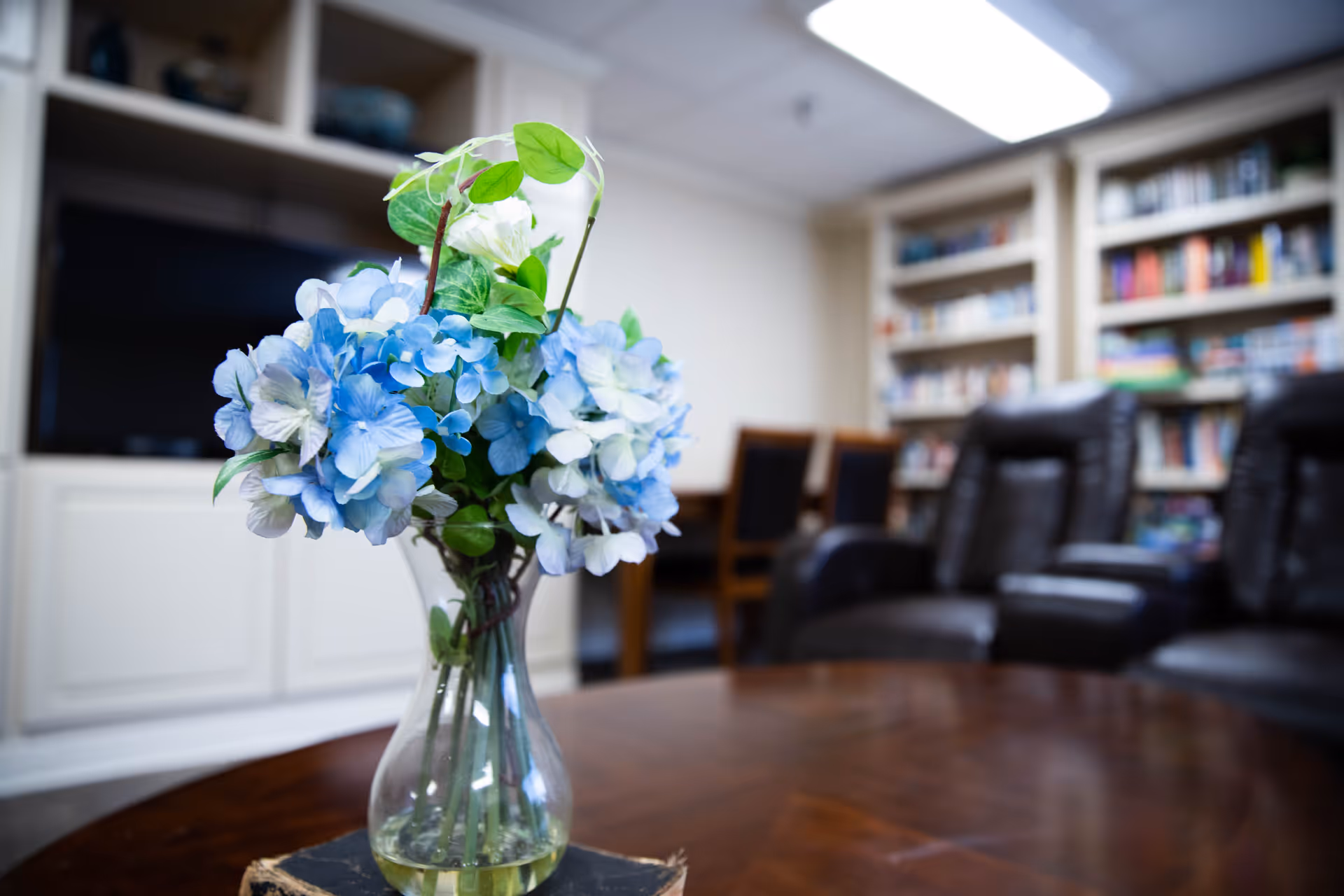 A clear glass vase with blue and white flowers sits on a wooden table in a cozy room. In the background, there are black leather chairs, a wooden table with chairs, built-in white shelves filled with books, and a television.