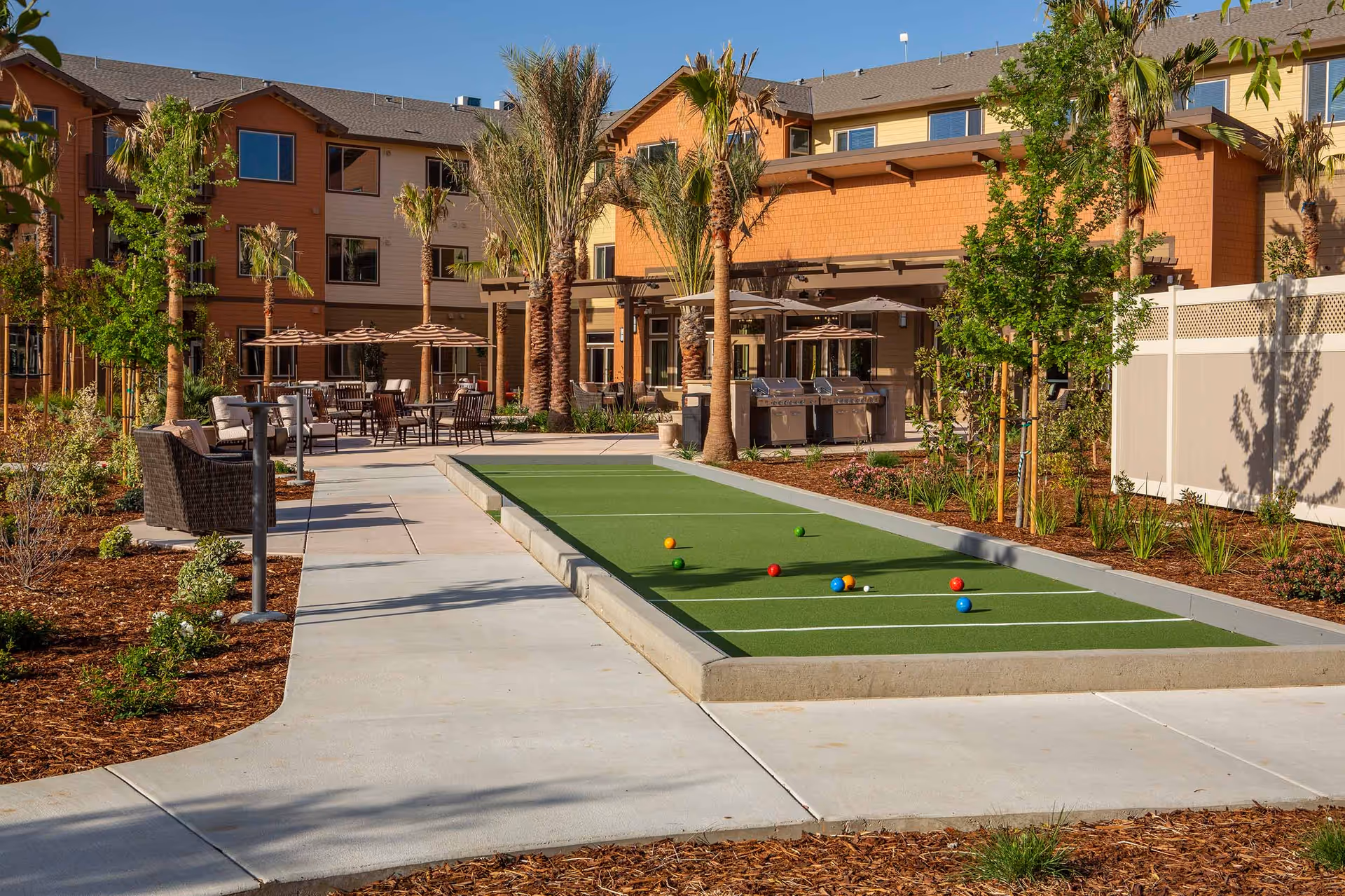 Outdoor recreational area at Bruceville Point featuring a bocce ball court with colorful balls, surrounded by palm trees and landscaped garden beds. There are seating areas with chairs and tables under umbrellas, and a building with multiple windows and a covered patio with grills in the background.
