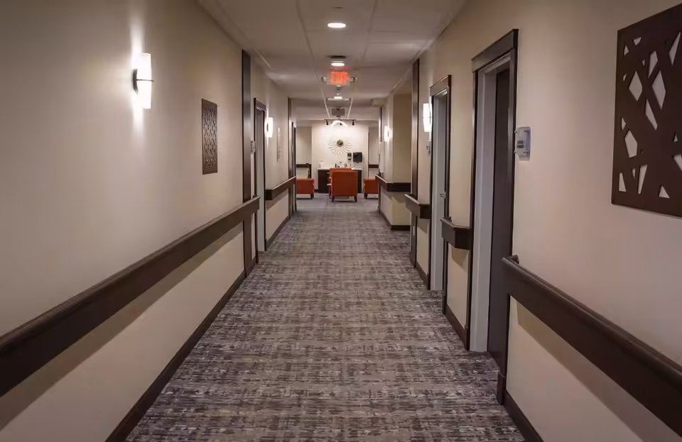 Carpeted hallway in a senior living facility with doors on both sides and a seating area at the far end.