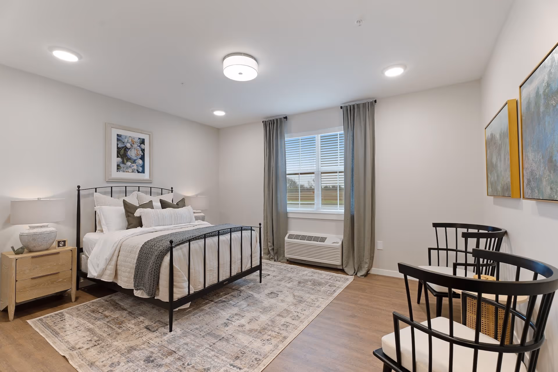 Bright, modern bedroom with a metal-framed bed, neutral bedding, nightstand, window with curtains, area rug, and two black chairs.