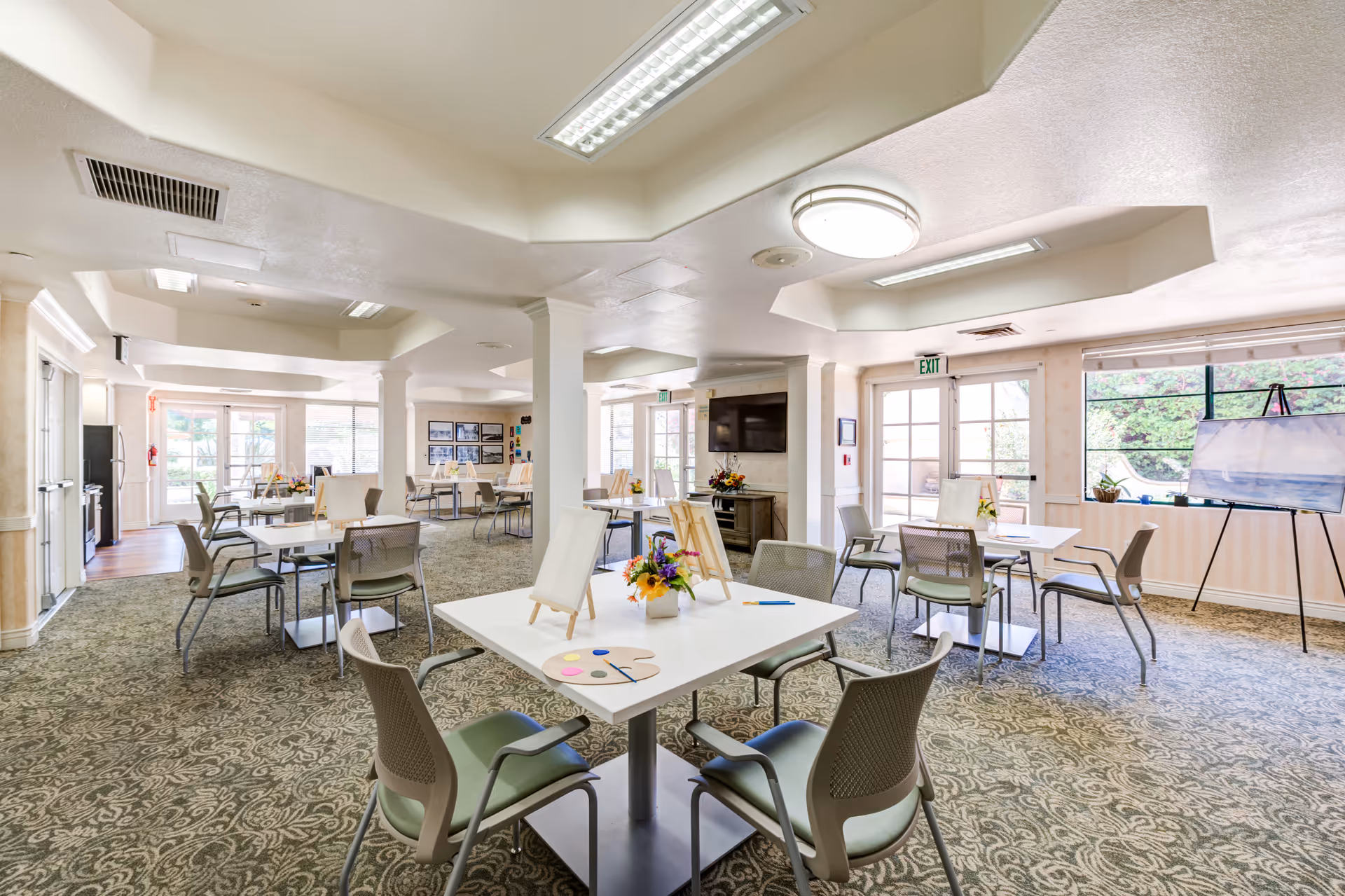 A spacious, well-lit activity room in a senior living facility with multiple tables and chairs arranged for group activities. Each table has small easels with blank canvases, paint palettes, and brushes, suggesting an art class setup. The room features large windows allowing natural light, patterned carpet flooring, and a television mounted on the wall. There are also floral centerpieces on the tables and an easel with a painting near the window.