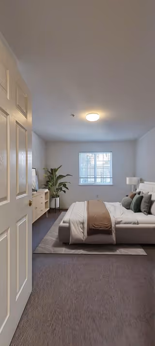 Bright furnished bedroom with a bed, nightstand, dresser, potted plant, and a window.