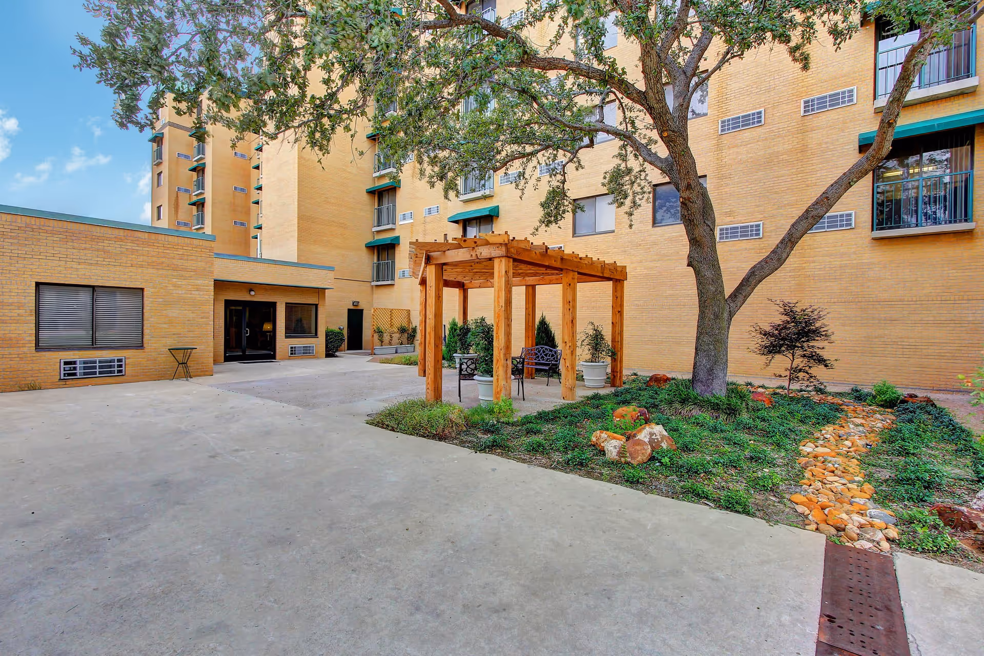 Outdoor courtyard area of a senior living facility with a wooden pergola, metal chairs, potted plants, a large tree, and a small landscaped garden with rocks and greenery. The building's yellow brick exterior and windows are visible in the background under a blue sky with some clouds.