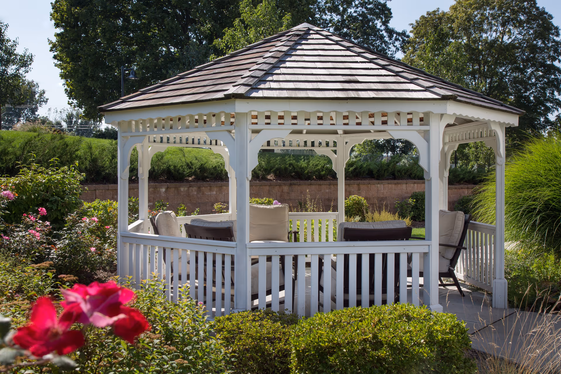 A white wooden gazebo with cushioned chairs inside, surrounded by green bushes, pink flowers, and trees under a clear sky.