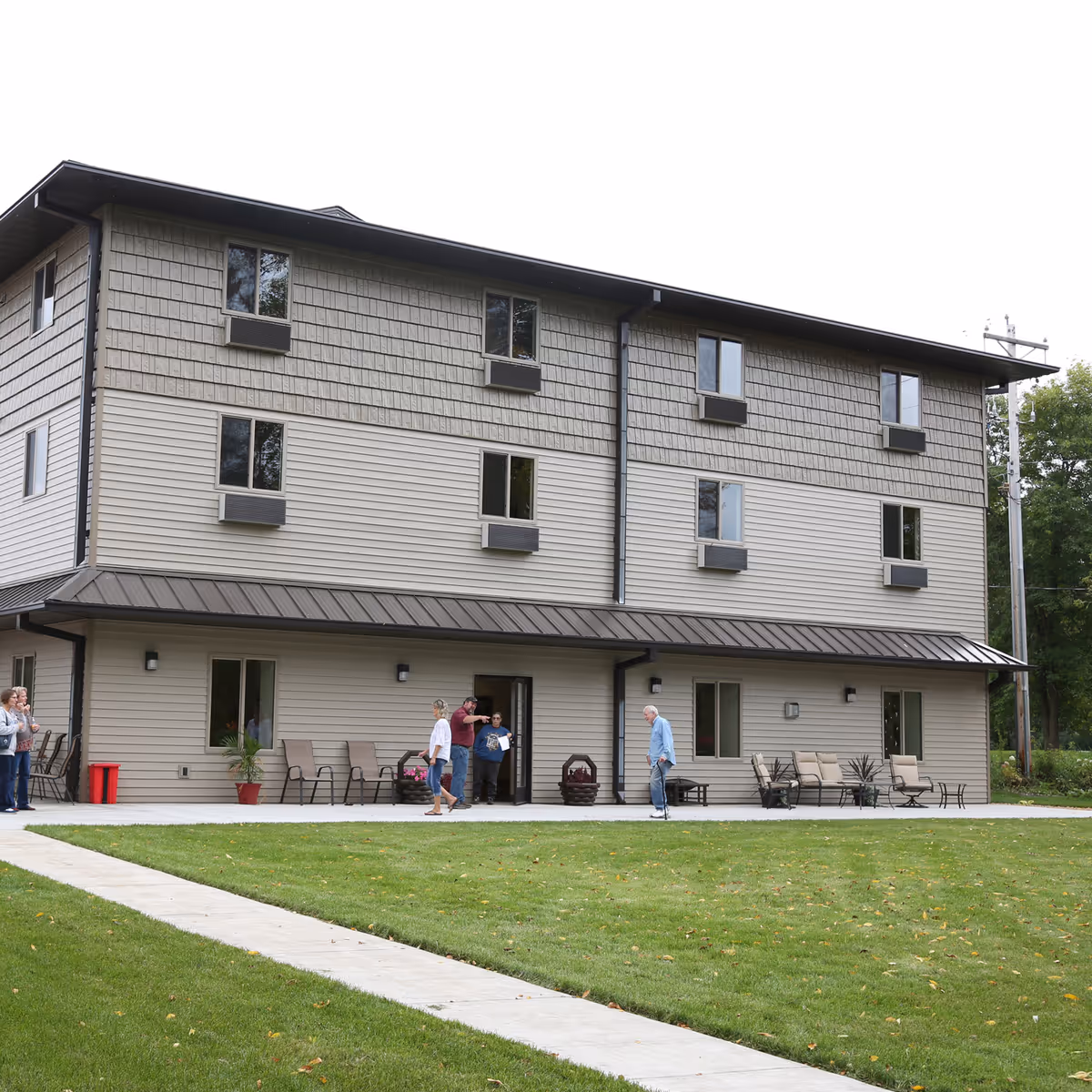 Exterior view of a three-story assisted living facility building with beige siding and multiple windows. Several people are standing and walking near the entrance on a paved patio area with outdoor chairs and tables. The building is surrounded by a well-maintained lawn and trees in the background.