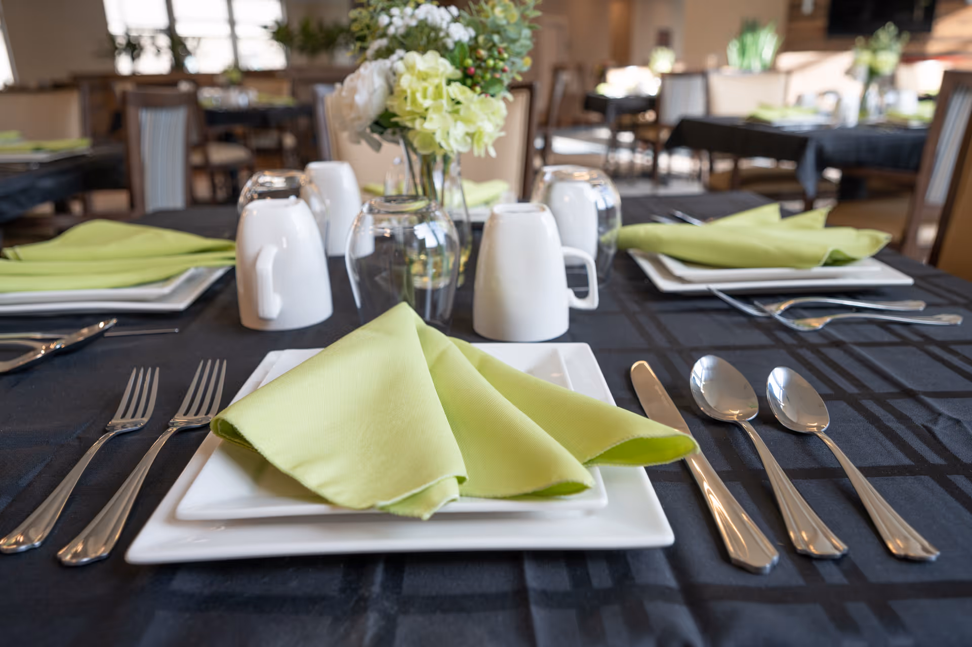 A close-up view of a dining table set with square white plates, green folded napkins, silverware including forks, knives, and spoons, upside-down white mugs, and clear glasses. In the center of the table is a small floral arrangement with white and green flowers. The tablecloth is black with a subtle checkered pattern, and other similarly set tables are visible in the background.