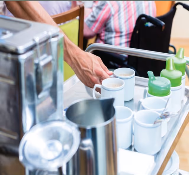 A person's hand placing coffee mugs and green-lidded sippy cups on a metal serving cart with pitchers and a coffee urn in a dining area.