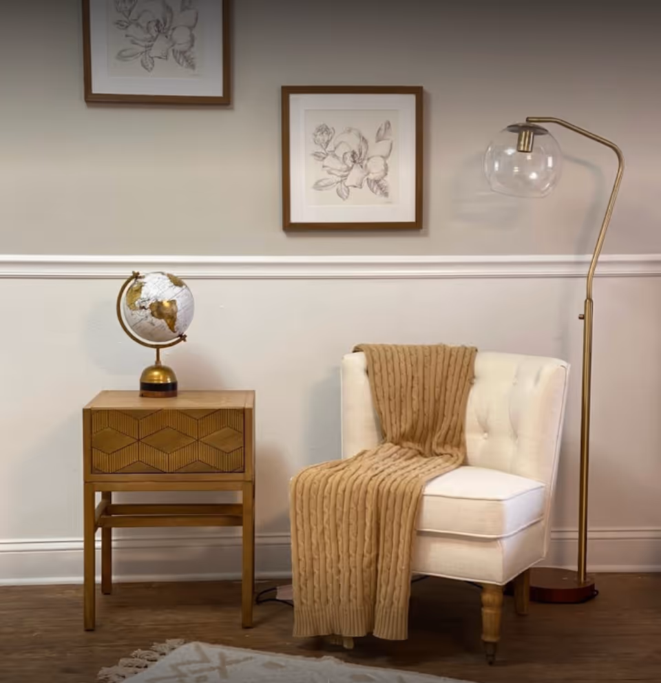 A cozy corner with a white upholstered chair draped with a tan knitted blanket, a wooden side table with a decorative globe on top, a tall floor lamp with a glass globe shade, and two framed floral sketches hanging on a beige wall.
