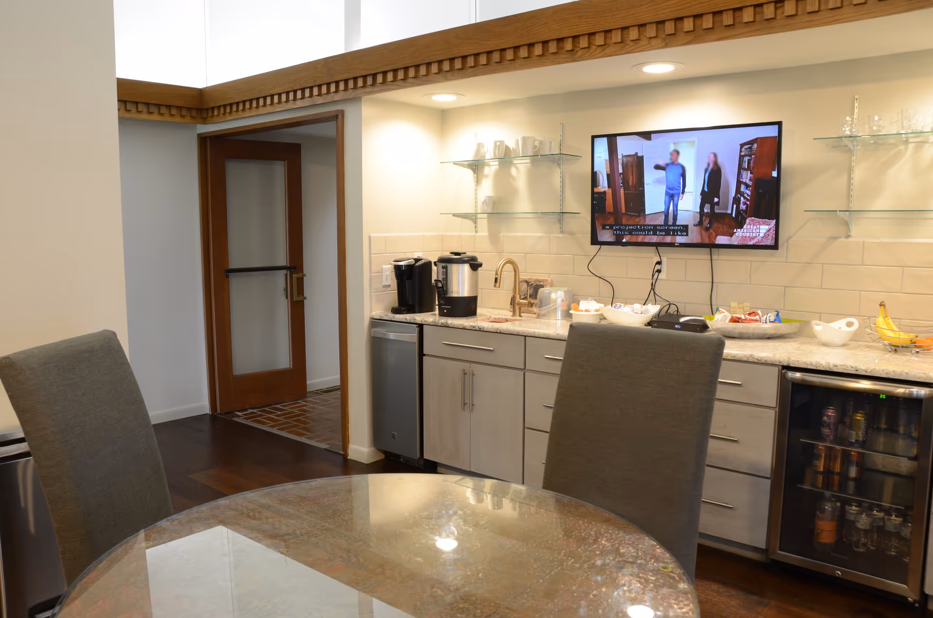 Interior view of a kitchen area in Southern Grace Assisted Living featuring a round glass table with two gray chairs, a countertop with a coffee maker, hot water dispenser, bowls of snacks, and a small refrigerator with beverages. A flat-screen TV is mounted on the wall above the counter, showing a program with two people. The room has light-colored walls, wooden flooring, and a door leading to another area.