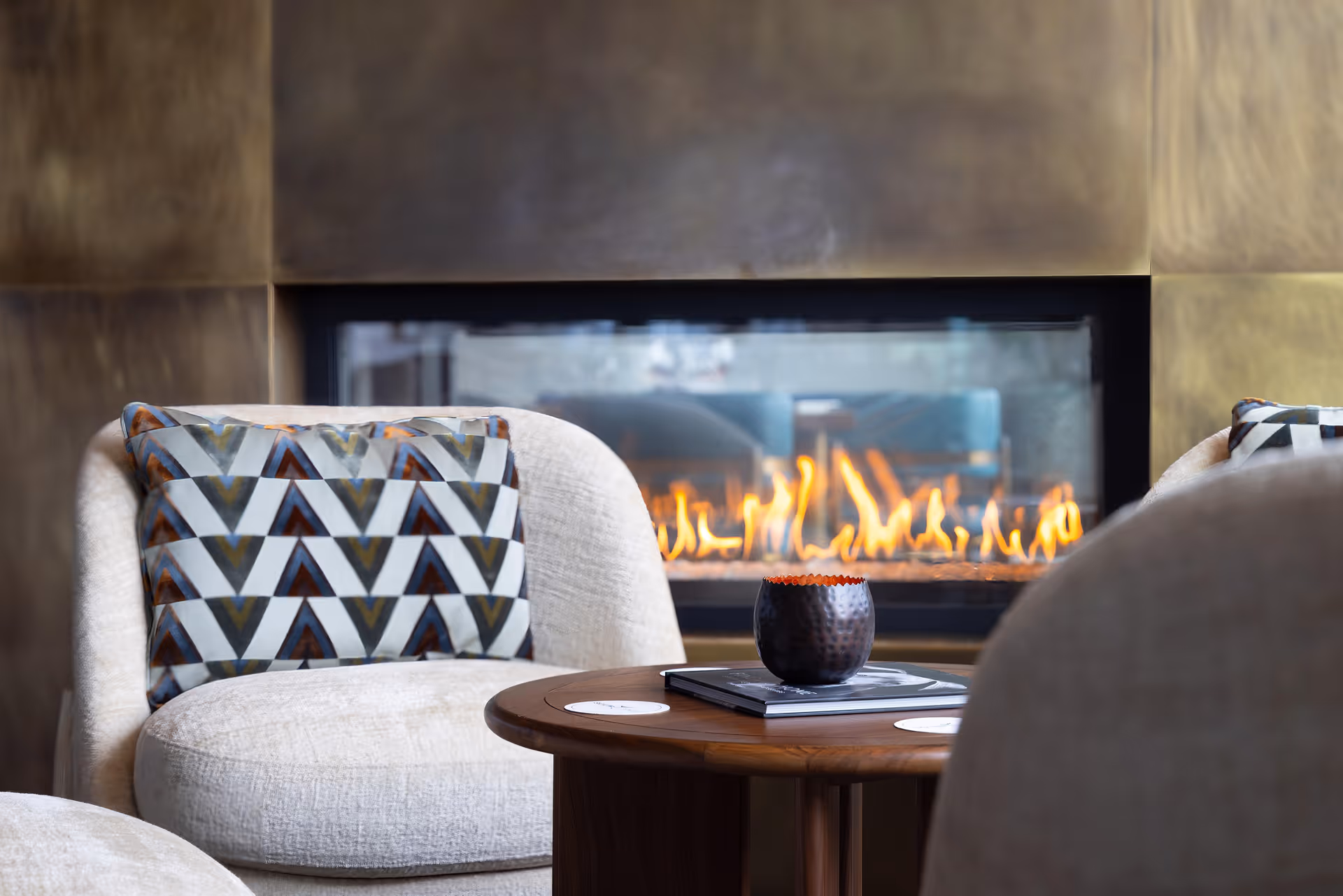 A cozy seating area with a modern fireplace in the background. The scene includes a round wooden table with a small decorative item and a book on it, surrounded by cushioned chairs with patterned pillows.