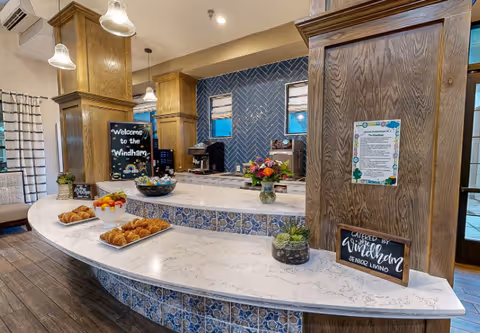 Interior view of a welcoming dining or serving area at The Windham senior living facility, featuring a curved white marble countertop with blue patterned tile on the front, plates of croissants and fruit, small potted plants, a coffee machine, and decorative signs including one that says 'Welcome to the Windham' and another that reads 'Catered by The Windham Senior Living'. The background includes wooden pillars, a blue herringbone tile wall, and windows with white blinds.