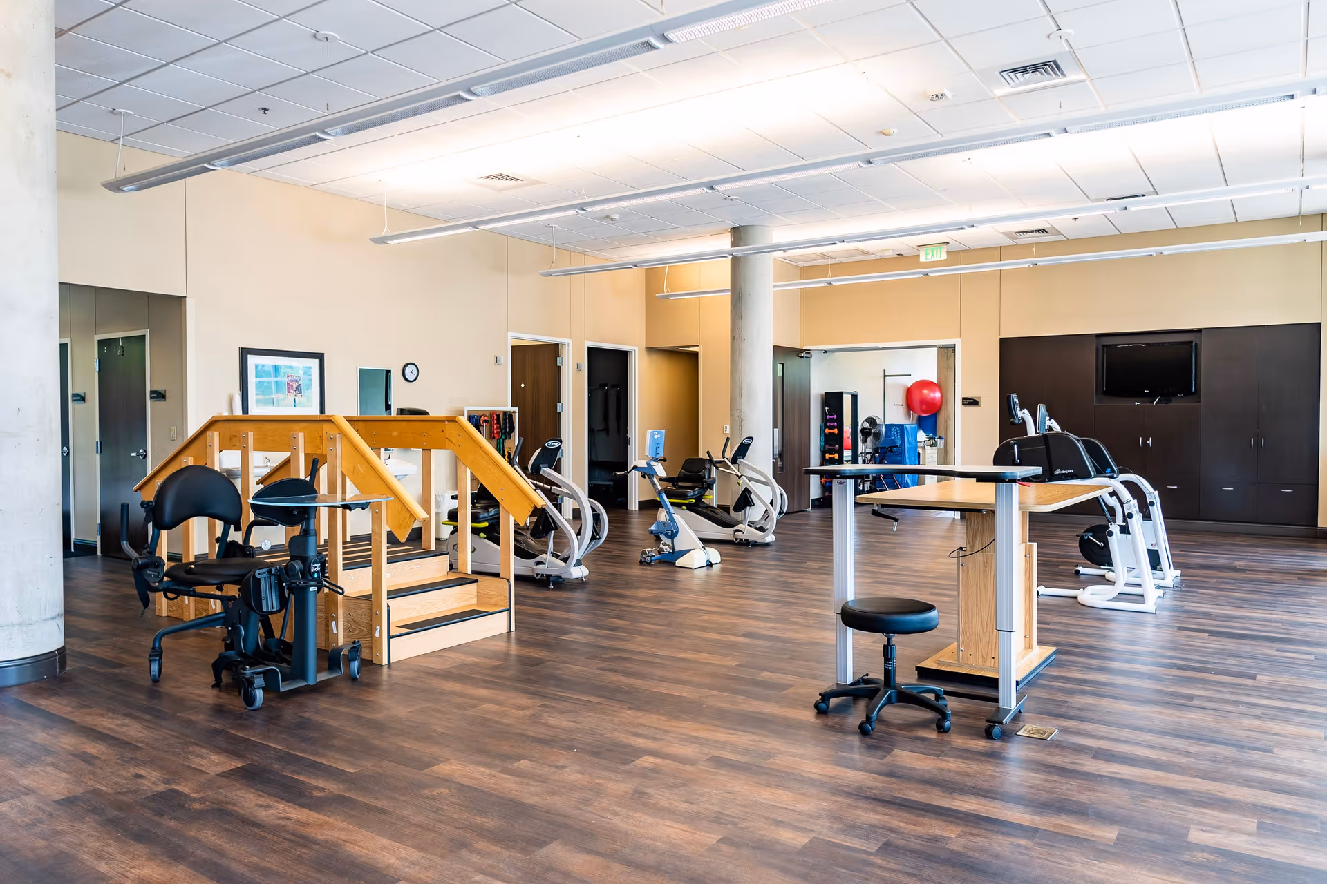 Spacious rehab gym with exercise machines, a wooden practice staircase, and seating in a bright interior room.