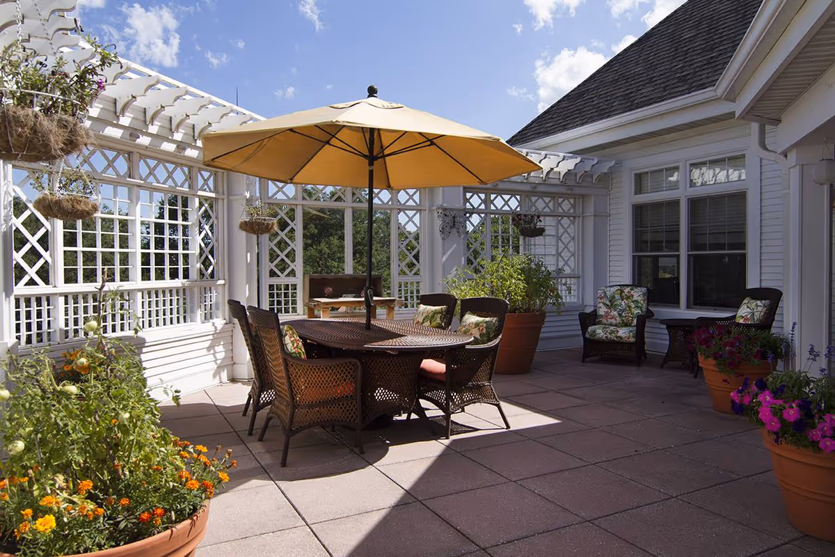 Outdoor patio area with a round table and six wicker chairs under a large yellow umbrella. The patio is surrounded by white lattice fencing and decorated with potted plants and flowers. There are two additional wicker chairs with floral cushions near the building wall with windows.