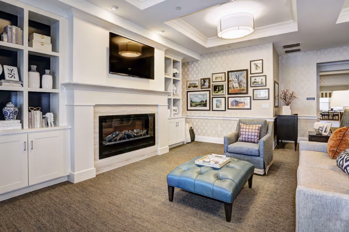 A cozy living room with a mounted flat-screen TV above a modern electric fireplace. Built-in white shelves and cabinets flank the fireplace, decorated with books, vases, and small sculptures. The room features a blue tufted ottoman, a gray armchair with a patterned pillow, and a beige sofa with orange and patterned cushions. The walls are adorned with framed artwork, and the ceiling has a recessed light fixture.