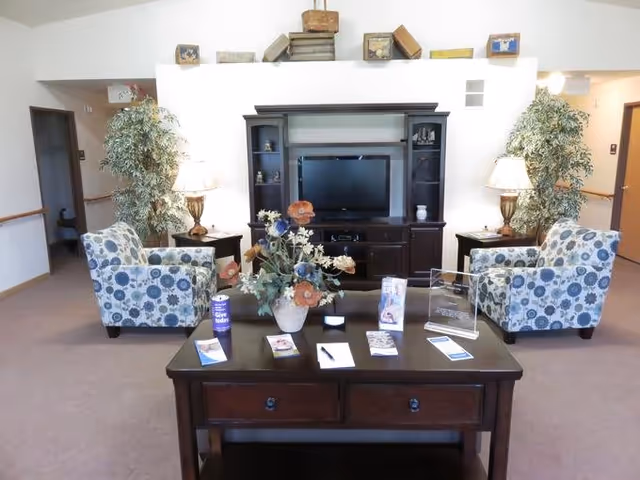 A cozy living room area in a senior living facility with two patterned armchairs facing a dark wood entertainment center holding a flat-screen TV. A wooden table in the foreground displays brochures, a pen, and a flower arrangement. Two table lamps and two large potted plants flank the entertainment center. The room has light-colored walls and carpeted floor.