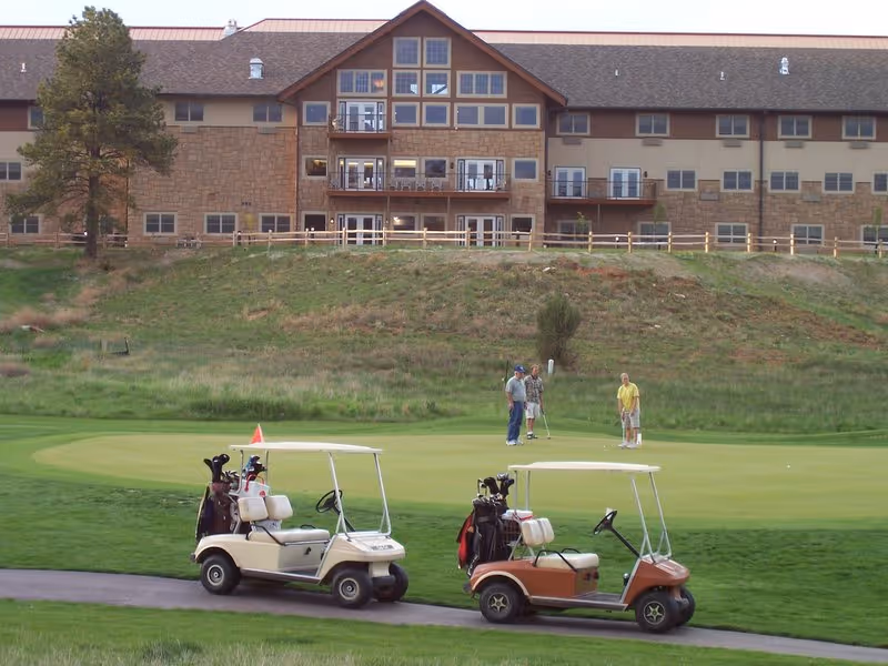 Two golf carts parked on a path near a putting green where three people are playing golf. In the background, there is a large multi-story building with many windows and balconies, set on a grassy hill.