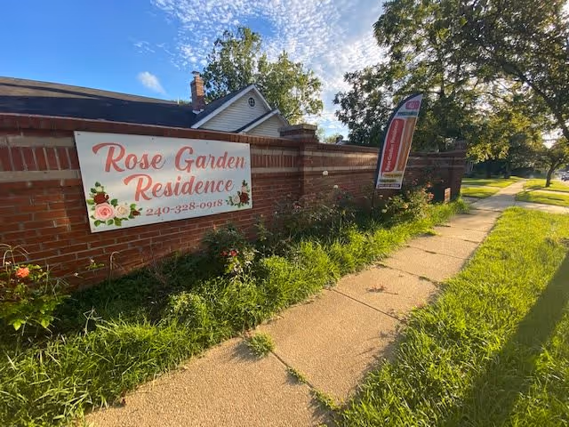 Brick perimeter wall beside a sidewalk with a banner and flag advertising 'Rose Garden Residence' amid grass and trees.