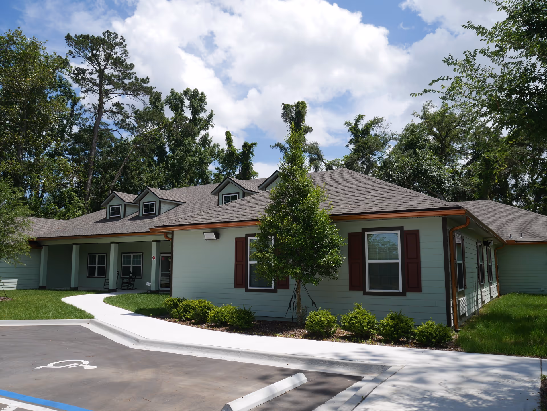 Single-story pale green residential building with dormer windows, a front walkway, landscaping and a parking area under a partly cloudy sky.