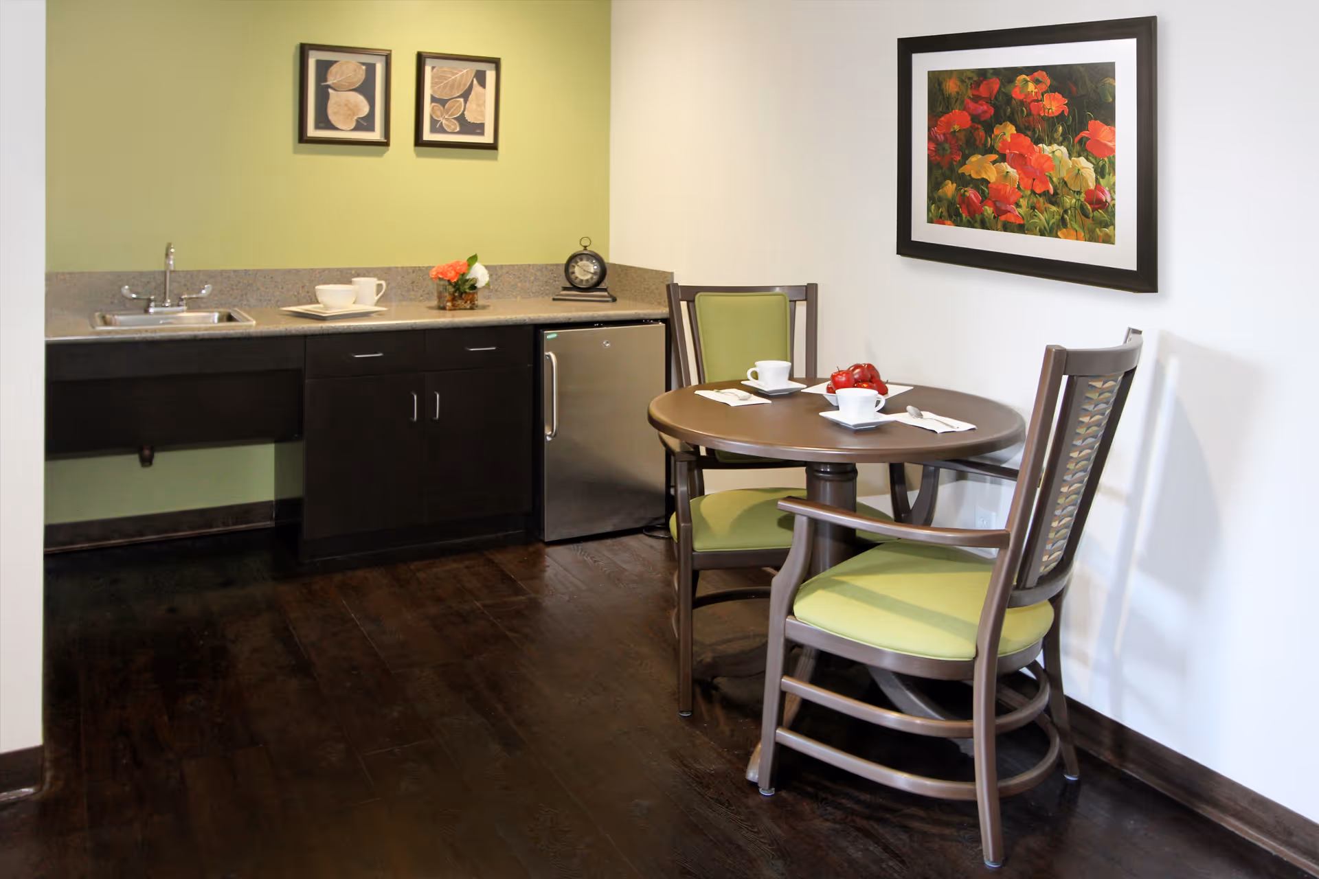 Small kitchenette area with a sink, countertop, and mini refrigerator. Next to it is a round wooden table with two chairs that have green cushions. On the table are two white cups and a bowl of red apples. The walls are decorated with two framed pictures, one with leaves and the other with red and yellow flowers.