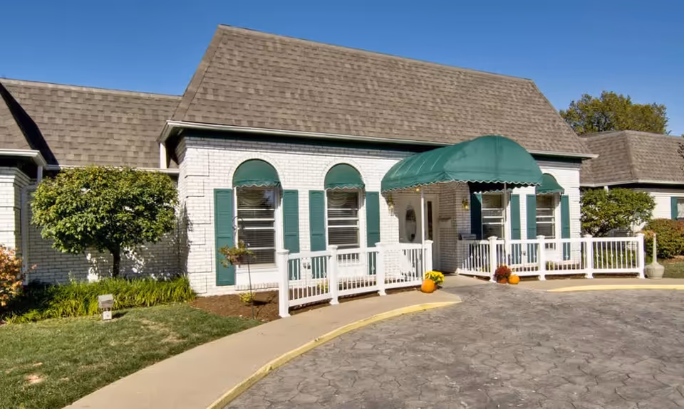 Exterior view of a single-story building with white brick walls and a brown shingled roof. The entrance features a green awning and white railings along a curved sidewalk. There are green shutters and arched windows, with some landscaping including a small tree and bushes. The sky is clear and blue.