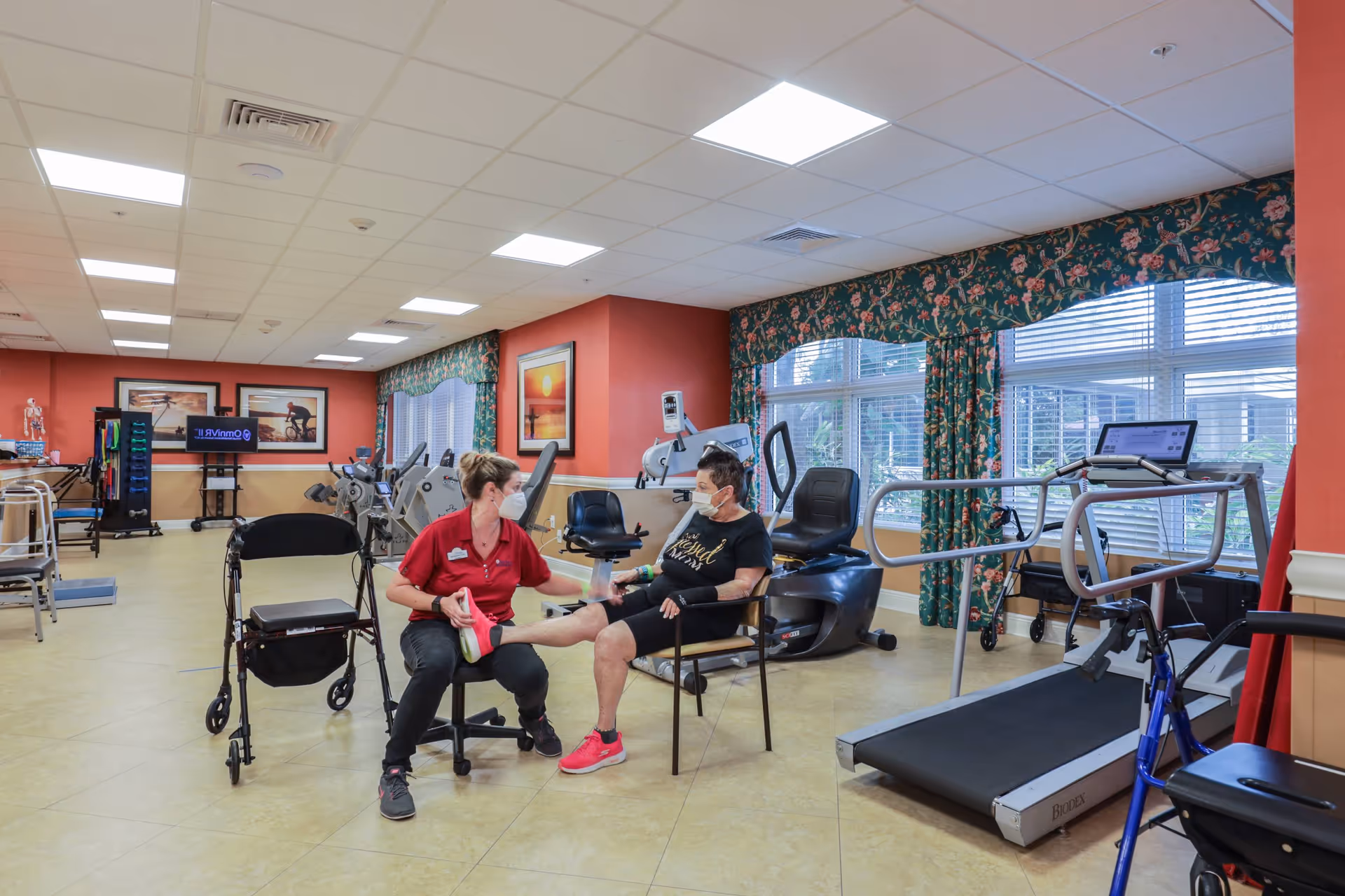 A physical therapy or exercise room in a senior living facility with exercise equipment including a treadmill, stationary bikes, and a walker. A staff member wearing a red shirt and face mask is assisting an elderly woman, also wearing a face mask, with a leg exercise while seated. The room has large windows with floral curtains and framed pictures on the walls.