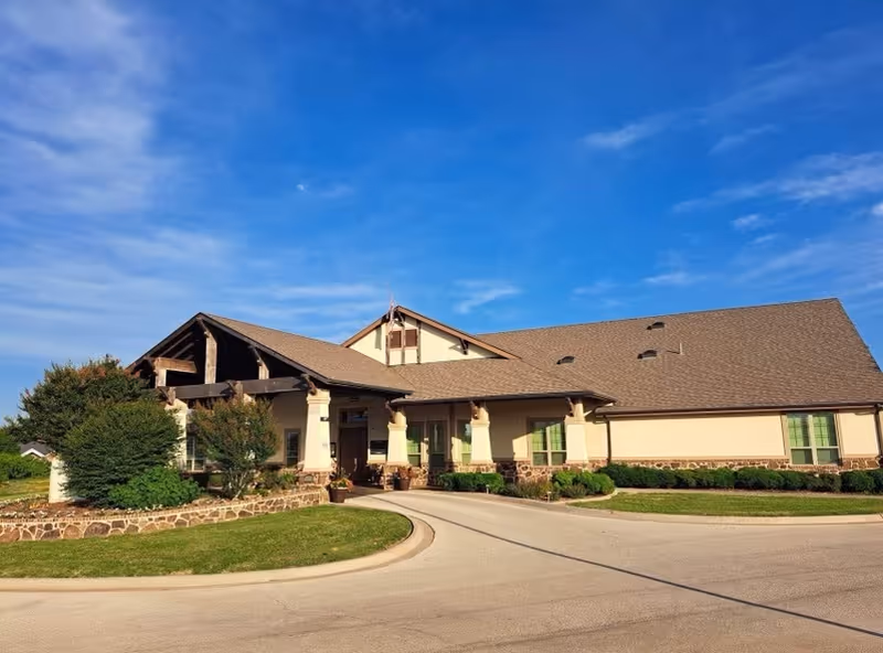 Exterior view of a single-story senior living facility building with a sloped roof, beige walls, stone accents, and green window shutters under a blue sky with some clouds. The entrance is covered by a porch supported by columns, and there is a curved driveway in front with landscaping including bushes and grass.