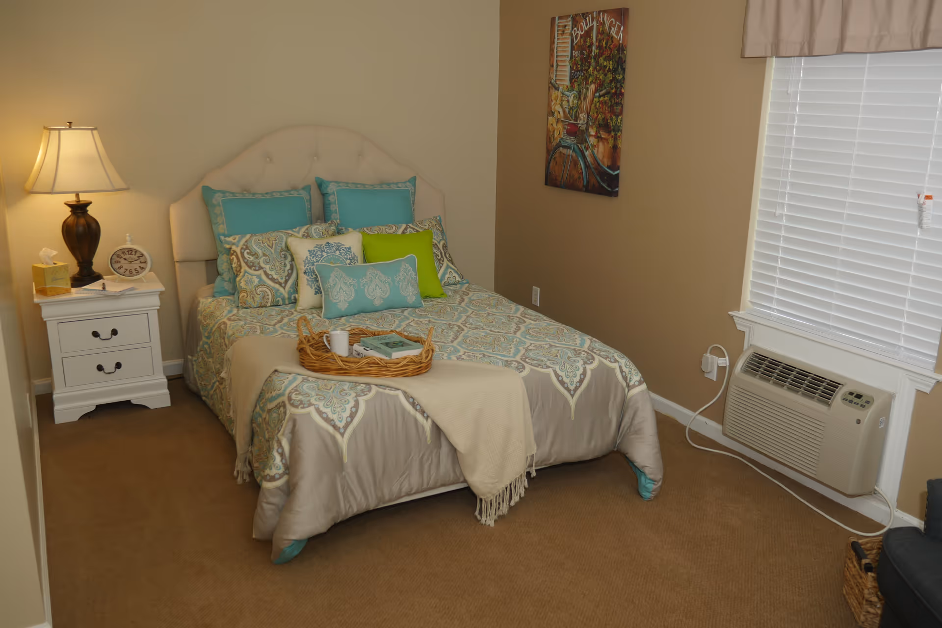 A furnished bedroom with a bed dressed in patterned linens, a nightstand and lamp, wall art, and a window air conditioning unit.