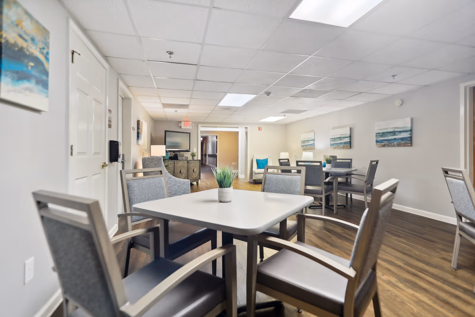 A bright and clean common area with several tables and chairs arranged for seating. The room has light-colored walls adorned with abstract ocean-themed paintings. There is a TV on a cabinet against the far wall and a comfortable armchair with a blue cushion near the back. The floor is wooden, and the ceiling has recessed lighting panels.