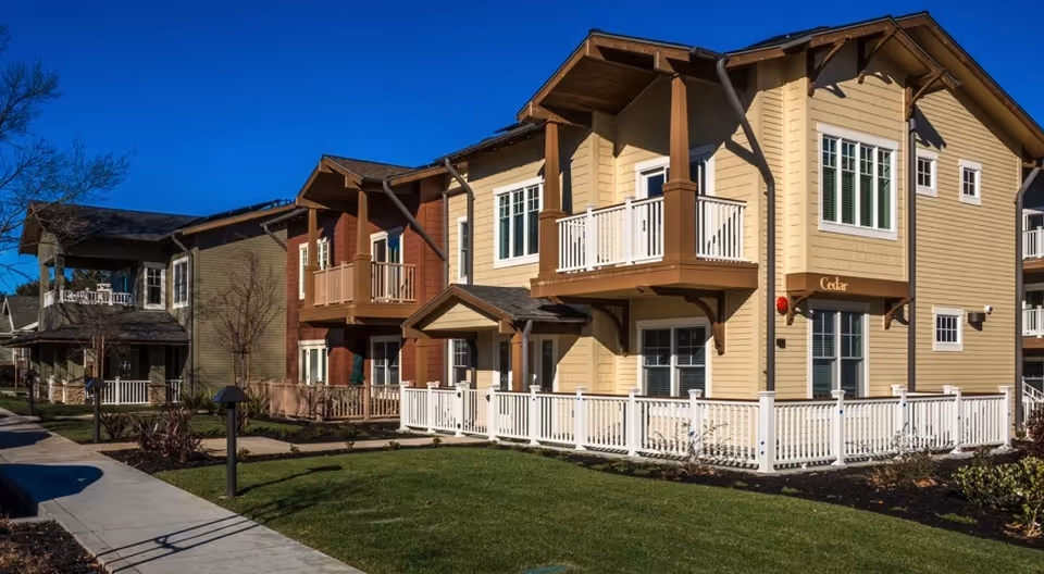Exterior view of a senior living facility building named Cedar at The Terraces at Los Altos, featuring beige and brown siding, white railings, balconies, and a well-maintained lawn under a clear blue sky.