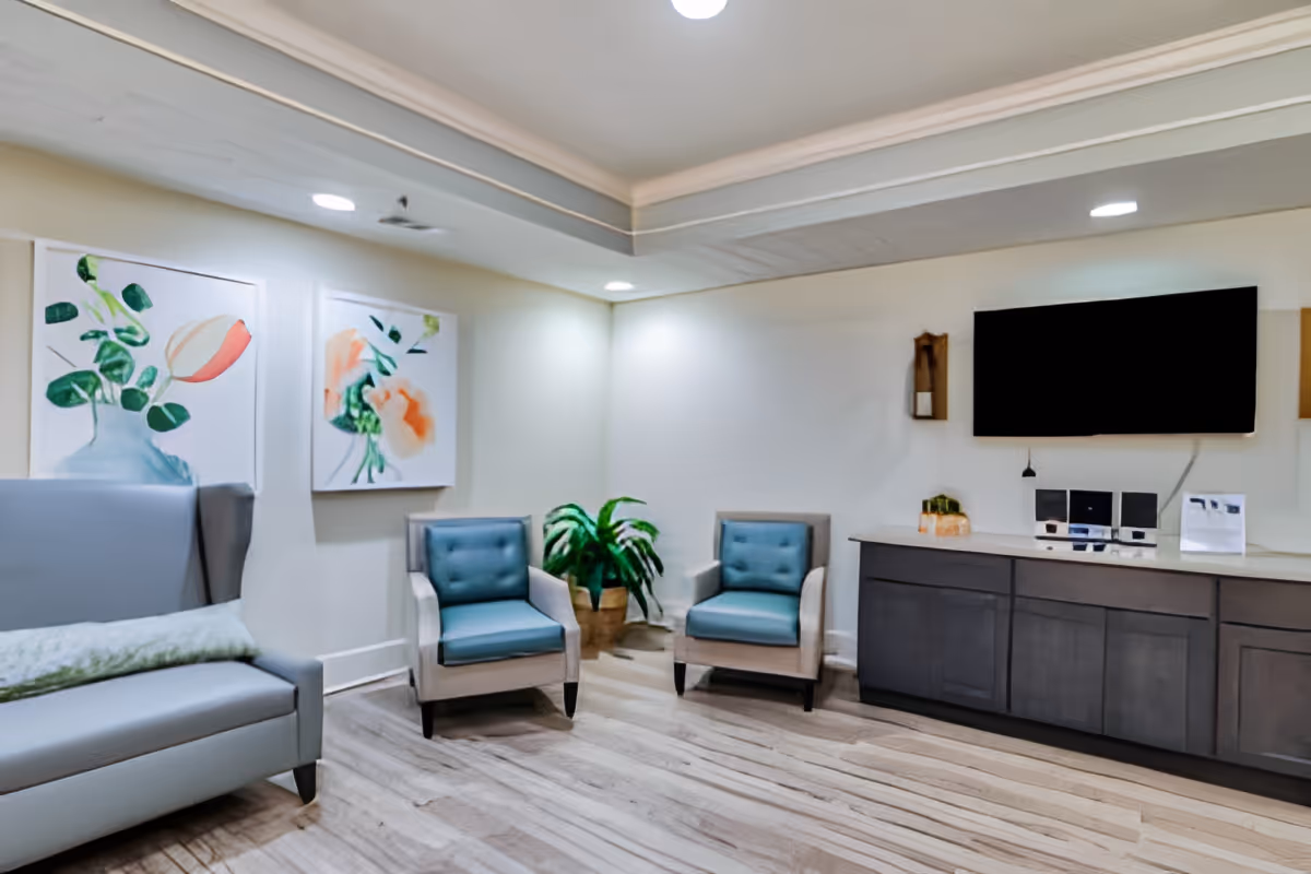 A bright lounge area with upholstered chairs and a sofa, wall art, potted plants, a TV mounted above cabinets, and wood-look flooring.