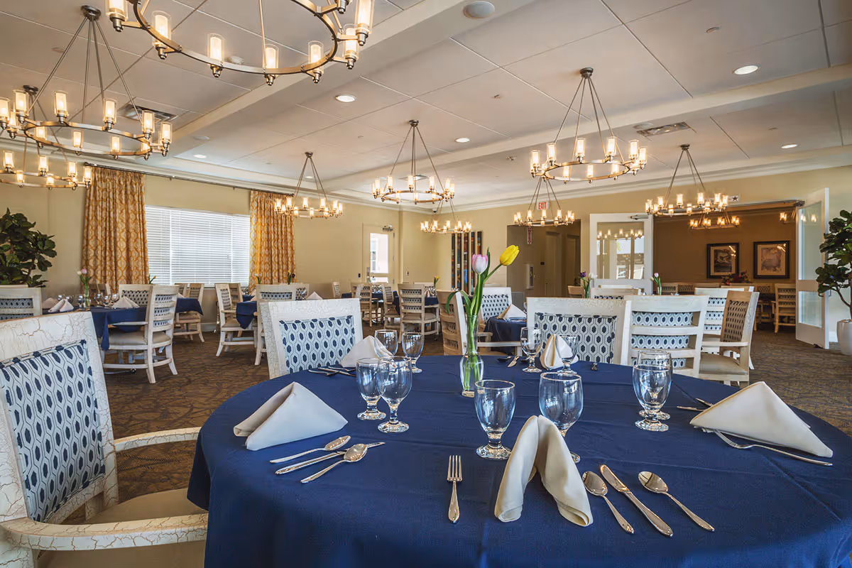 Elegant dining room with round tables covered in blue tablecloths, place settings, and hanging chandeliers.