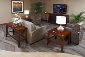 A cozy living room area with a beige sofa, two patterned armchairs, two wooden side tables with lamps, a wooden TV stand with a flat-screen TV, a large potted plant, and a framed picture on the wall.