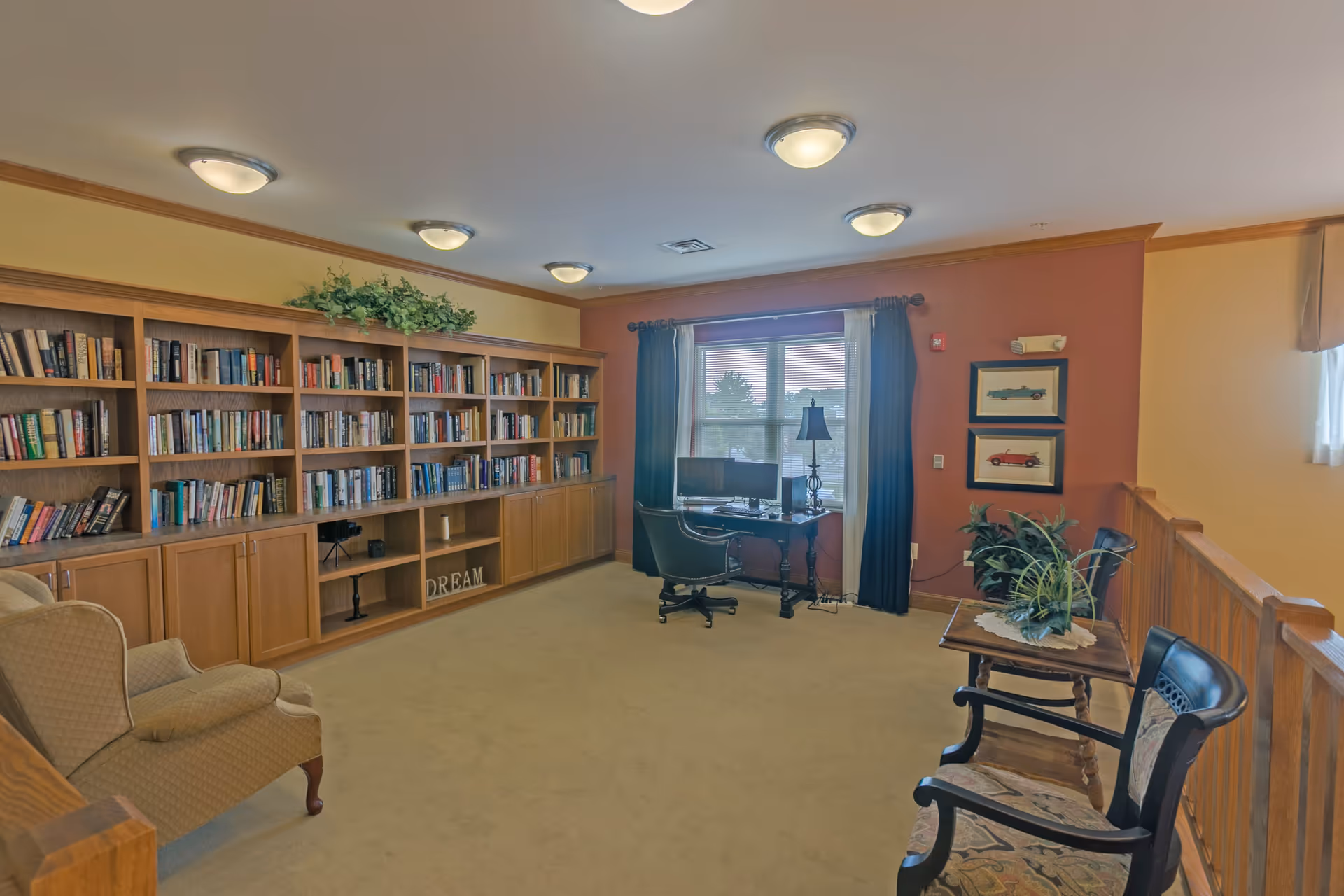 A cozy interior room with a large wooden bookshelf filled with books along one wall, a beige armchair, a desk with a computer and chair near a window with curtains, two framed pictures of vintage cars on a red accent wall, and two wooden chairs with a small table holding a plant.
